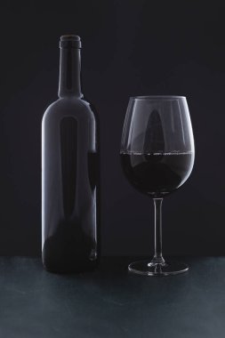 Yeşil ahşap bir yüzey üzerinde, siyah bir arkaplanın önünde etiketsiz bir şişe kırmızı şarap ve dolu bir bardak var..