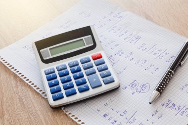 Tahta bir masanın üzerinde el yazısıyla yazılmış sayılar ve hesaplamalarla dolu bir defter karesi var. Kağıdın üzerinde de kalem ve hesap makinesi var..