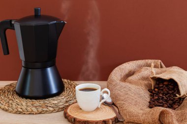 Bir masanın üzerinde yuvarlak bir hasır masa örtüsünün üzerinde siyah bir kahve demliği, sıcak kahve ile beyaz bir porselen bardak ve küçük bir çuvaldan çıkan bir avuç kahve çekirdeği var..