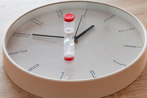 A large round needle clock there is a white hourglass with red ends.The clock strikes ten to two.