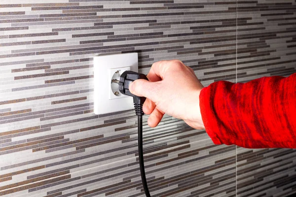 Bilinmeyen bir adamın eli, karo bir duvara takılmış bir elektrik prizine siyah bir Avrupa prizi takıyor. Elbisenin kolu kırmızı..