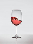 Křišťálová sklenice s růžovým vínem v pohybu. Noha skla se odráží na povrchu a pozadí je bílé.