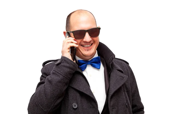 Kel, beyaz, şık giyinmiş ve güneş gözlüğü takan bir erkek gülümserken cep telefonuyla konuşuyor. Arkaplan beyaz.