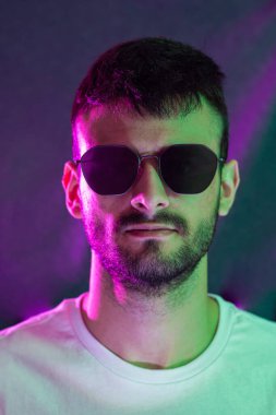 Sakallı, güneş gözlüğü takan, yeşil ve morumsu ışıklarla aydınlatılmış kameraya bakan beyaz bir adamın portresi..