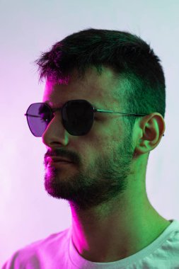 Sakallı, güneş gözlüğü takan, yeşil ve morumsu ışıklarla aydınlatılmış beyaz bir adamın portresi..