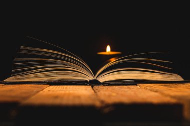 Yarı karanlıkta, arka planda yanan bir mumla birlikte antik ahşap bir masa üzerinde açık bir kitabın sayfalarının kenarlarını kapatın..