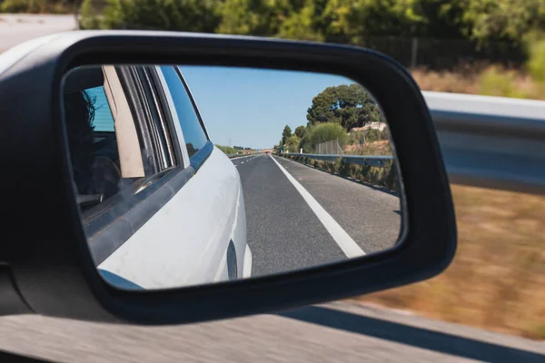 Rearview mirror of a white car driving on a highway, which is reflected in the mirror. In the background you can see the sweeping movement of the exterior.