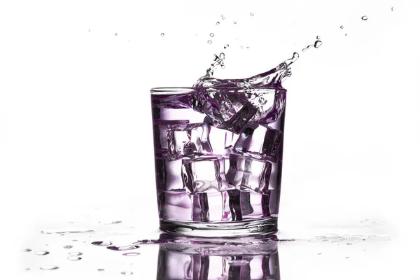A crystal glass with ice and a purple liquid splashing with the fall of an ice cube, isolated on a white background and reflected on the surface.