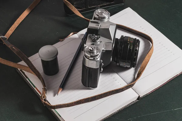 An antique analog camera on an diary in which there is a pencil and a bottle of photographic film. There is a strap and a leather case.