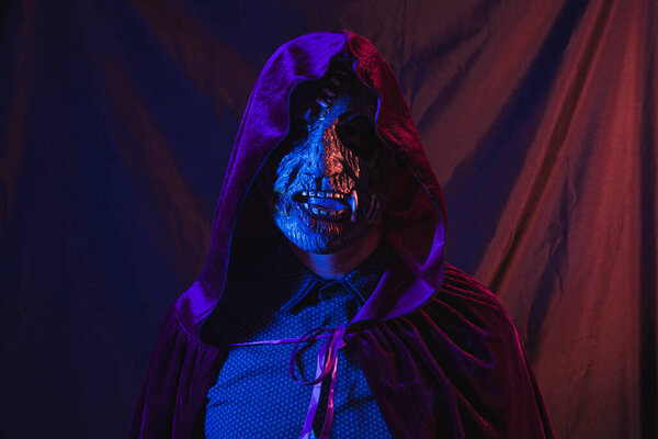 Portrait of a zombie dressed in a shirt and hooded cape facing the camera. The scene is dark, illuminated by blue and orange lights.