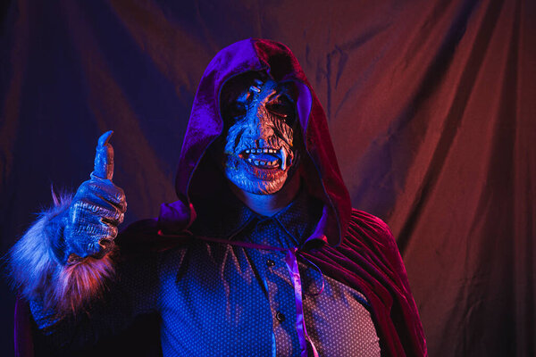 Portrait of a zombie dressed in a shirt and hooded cape making the like gesture with thumbs up. The scene is dark, illuminated by blue and orange lights.