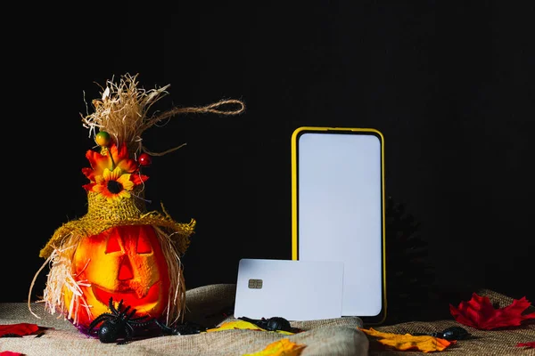 Credit card on a cell phone with a yellow case and the screen on, both standing next to an ornate Halloween pumpkin, on sackcloth and surrounded by toy spiders and fallen leaves next to pine cones.