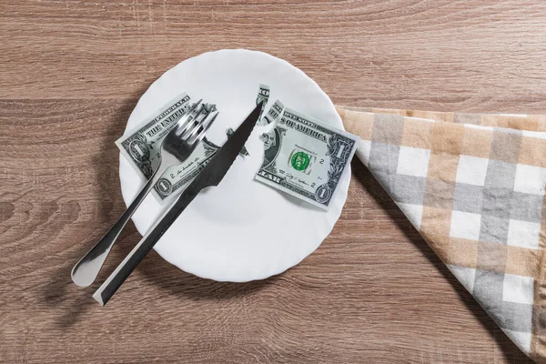 A white plate with a dollar bill torn into two large pieces and a few small quarters. On the plate is a knife and fork. It is on a wooden table and next to it is a cloth napkin.