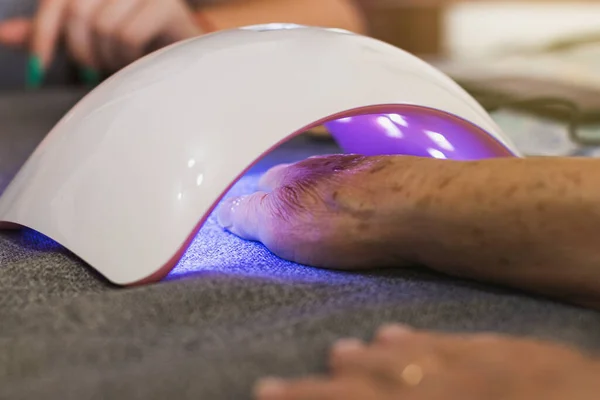 Close-up of an unrecognizable older woman's hand tucked under a gel nail dryer with a purple light.