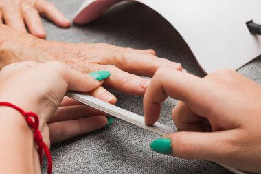 Close-up of the hands of two unrecognizable women, one young and one old, having a manicure. The young woman is filing the older woman's nails with a nail file.