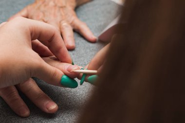Close-up of the hands of two unrecognizable women, one young and one old, having a manicure. The young woman, with long hair, is cleaning the old woman's nails with a wooden stick.