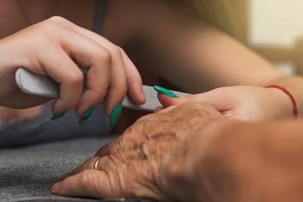 Close-up of the hands of two unrecognizable women, one young and one old, having a manicure. The young woman is filing the front of the older woman's nails with a nail file.