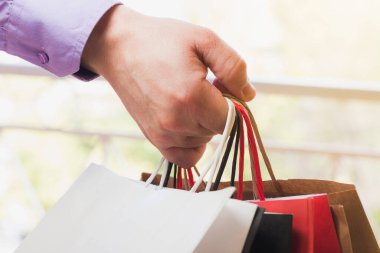 Close-up of the hand of an unrecognizable Caucasian man dressed in a purple shirt holding different colored paper shopping bags on the street.