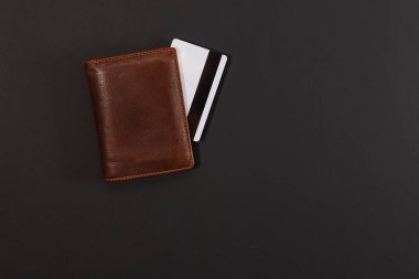 A brown leather wallet and a white credit card seen from the magnetic stripe side isolated on a dark background. On one side there is empty space.