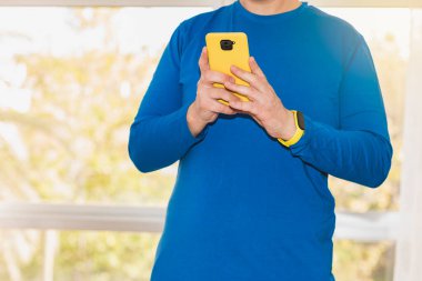 Mavi, uzun kollu tişört giymiş beyaz bir adam sarı renkli çantalı bir akıllı telefon kullanıyor. Aktivite bilekliği takıyor. Arka planda bir pencere var..