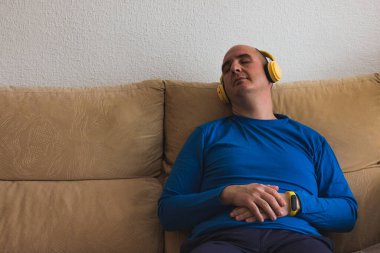 Uzun kollu mavi tişört giymiş kel bir beyaz adam, sarı kulaklık takmış, koltukta uyuyor..