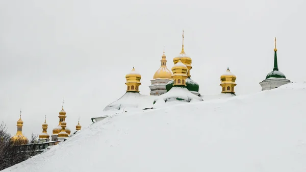 Golden domes of Kiev Pechersk Lavra or Kyiv Monastery of Caves behind a snowy slope on cold winter day. Cupolas of famous orthodox church on Dnipro river bank, Ukraine — Stock Photo, Image
