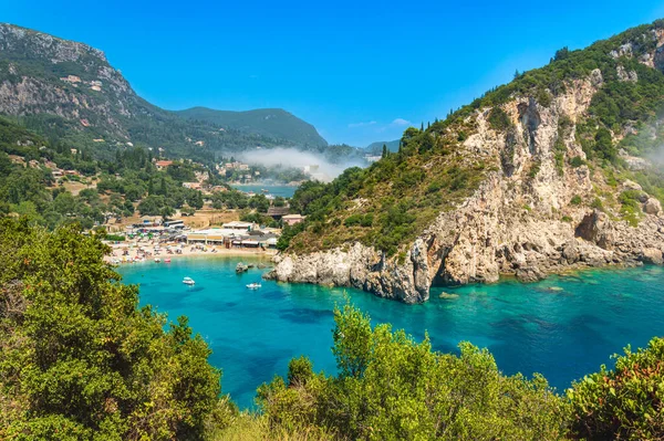 Paleokastritsa playa y pueblo en la isla de Corfú, Grecia. Pintoresca orilla del mar con agua cristalina turquesa, playa de guijarros Agios Spiridon, montañas y acantilados — Foto de Stock