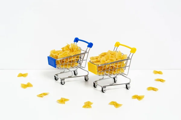 Uncooked farfalle pasta in blue and yellow shopping carts in colors of Ukrainian flag isolated on white background. Food supply crisis, price increase for wheat products, groceries shopping concept — Stock fotografie