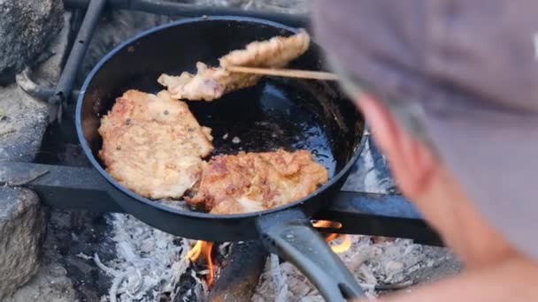 Человек готовит жареную свиную отбивную в масле на сковородке над огнем, жирное мясо — стоковое видео