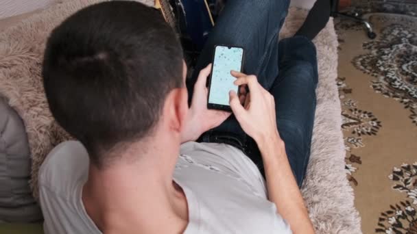 Человек смотрит на карту с отмеченными точками на смартфоне, пока лежит на диване в комнате — стоковое видео