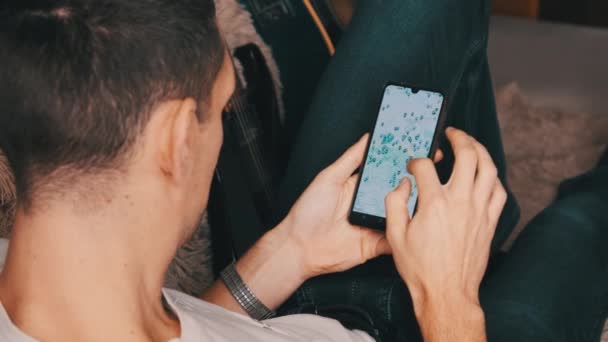 Homem olhando para o mapa com pontos marcados no smartphone enquanto deitado no sofá no quarto — Vídeo de Stock