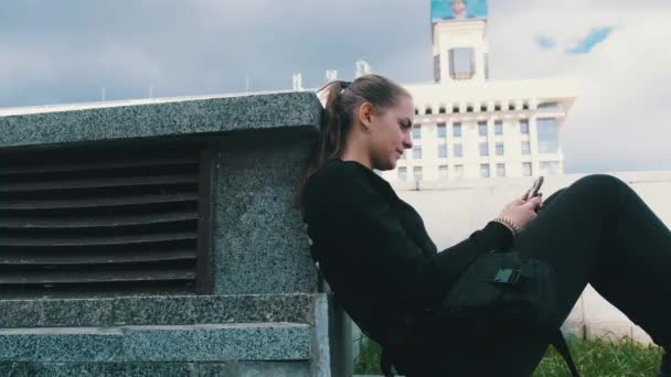 Mujer joven utiliza Smartphone mientras está sentado en el parapeto en el parque de la calle — Vídeos de Stock