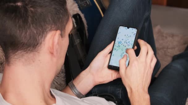 Человек смотрит на карту с отмеченными точками на смартфоне, пока лежит на диване в комнате — стоковое видео