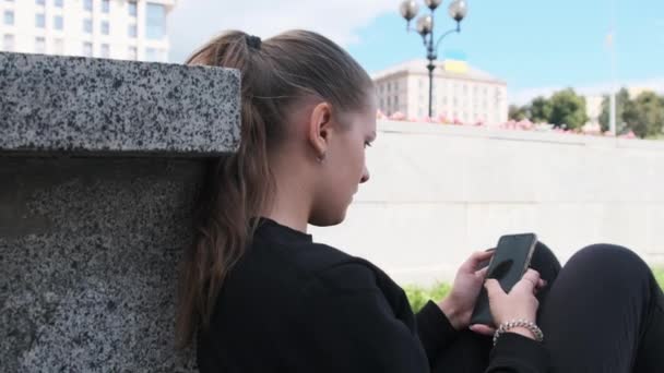 Молодая женщина пользуется смартфоном, сидя на парапете в парке — стоковое видео