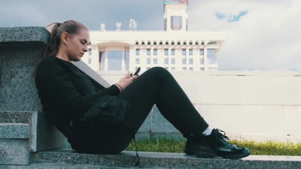 Молодая женщина пользуется смартфоном, сидя на парапете в парке — стоковое видео