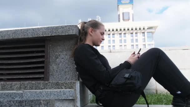Mujer joven utiliza Smartphone mientras está sentado en el parapeto en el parque de la calle — Vídeos de Stock