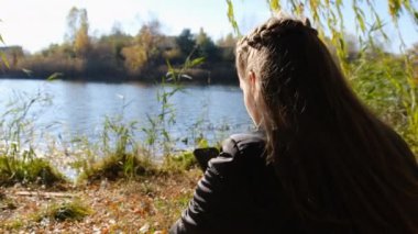 Ellerinde Akıllı Telefonlu Genç Kadın Sonbaharda Nehir kenarındaki Sarı Yeşillikte Oturuyor