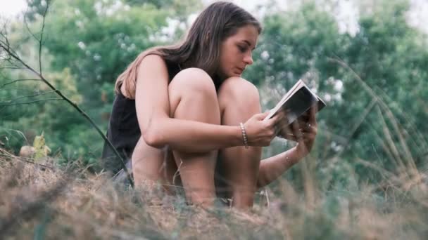 Молодая женщина читает книгу, сидя на траве в природе в летний день — стоковое видео