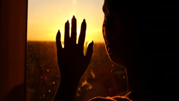 Mulher silhueta na janela colocando a mão no vidro através de raios de pôr do sol câmera lenta — Vídeo de Stock