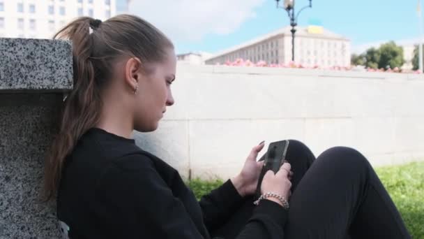 Jovem mulher usa smartphone enquanto sentado no parapeito no parque de rua — Vídeo de Stock