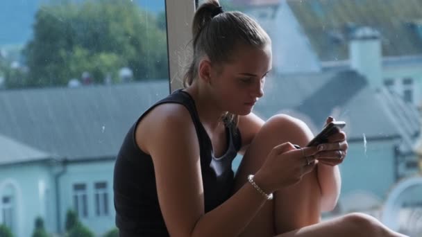 スマートフォンを持つかなり若い女性は、都市の窓のそばに窓の上に座る — ストック動画