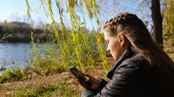 秋には手にスマートフォンを持った若い女性が川のそばで黄色の葉に座る — ストック動画