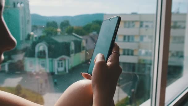 Красивая молодая женщина со смартфоном сидит на подоконнике у окна в городе — стоковое видео