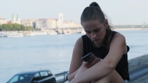 Молодая женщина пользуется смартфоном на набережной в парке возле реки — стоковое видео