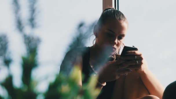 Mujer bastante joven con Smartphone se sienta en el alféizar de la ventana en la ciudad — Vídeo de stock