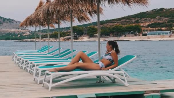 Aantrekkelijke vrouw zonnebaden op dek stoel onder stro paraplu door turquoise zee — Stockvideo