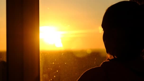 Silueta de una mujer joven mirando por la ventana al atardecer, cámara lenta — Vídeos de Stock