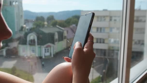 Досить молода жінка зі смартфоном сидить на підвіконні біля вікна в місті — стокове відео
