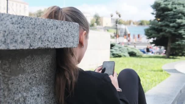 Mujer joven utiliza Smartphone mientras está sentado en el parapeto en el parque de la calle — Vídeos de Stock