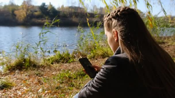 손에 스마트폰을 들고 있는 젊은 여인, 가을에 강가에서 옐로 포즈를 취하다 — 비디오